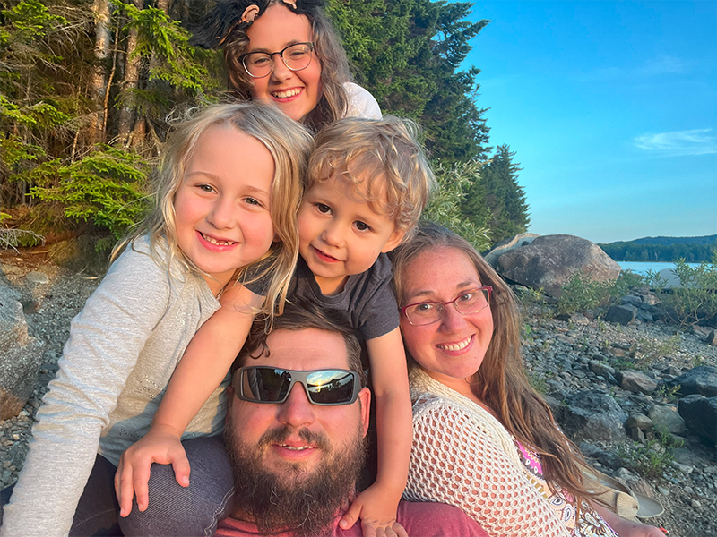 Alt Text
            Sarah with her husband Peter and all three children, clustered together on the shores of a rocky lake, with pine trees, boulders, and the lake in the background 
            
            