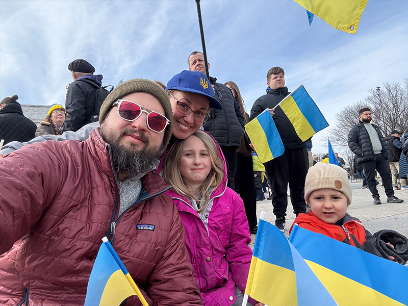 Sarah, her husband, two of their children, in winter jackets on the steps of the national mall, with Ukrainian flags. Sarah is wearing a blue hat with a yellow Ukrainian trident symbol. 