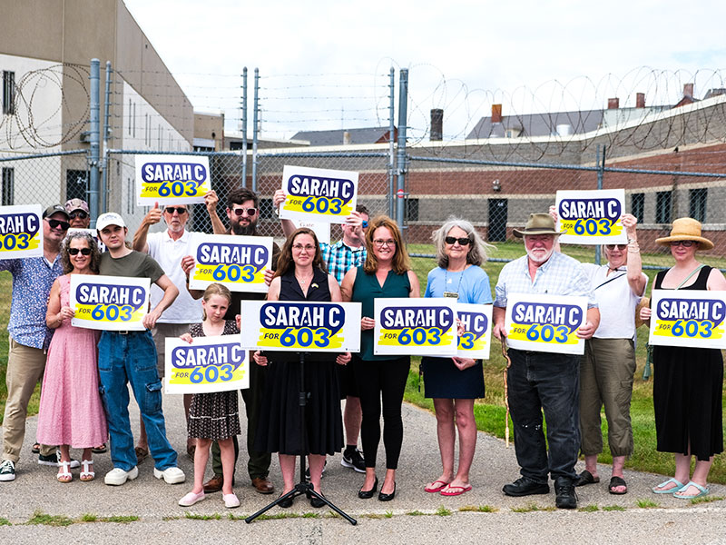 Sarah standing at a black podium, wearing a black dress,with a sign that is a mix of blue and yellow, that reads SarahC for 603, flanked by a larger group of people all holding signs that read SarahC for 603, there is green grass in the background broken by a tall metal fence with barbed wire on the top, and a brick building in the background 